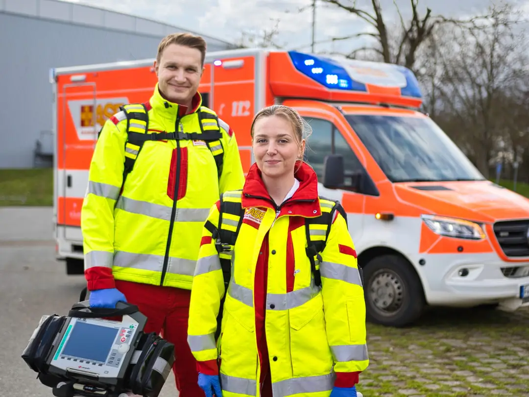 Zwei Kolleg:innen des ASB-Rettungsdienstes mit Notfallrucksack und Defibrillator vor einem Rettungswagen.